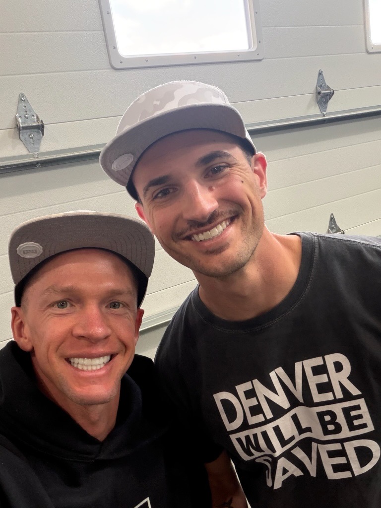 Close-up of two smiling men, one wearing the Denver Will Be Saved t-shirt
