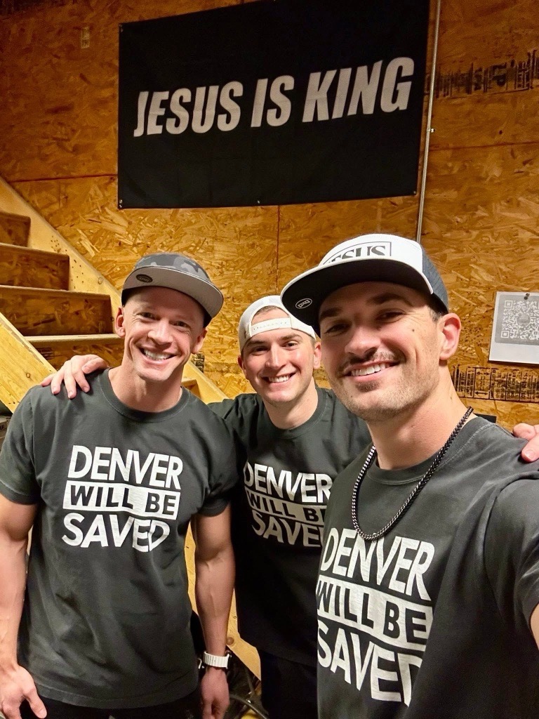 Three men posing together in matching Denver Will Be Saved shirts under a Jesus Is King banner